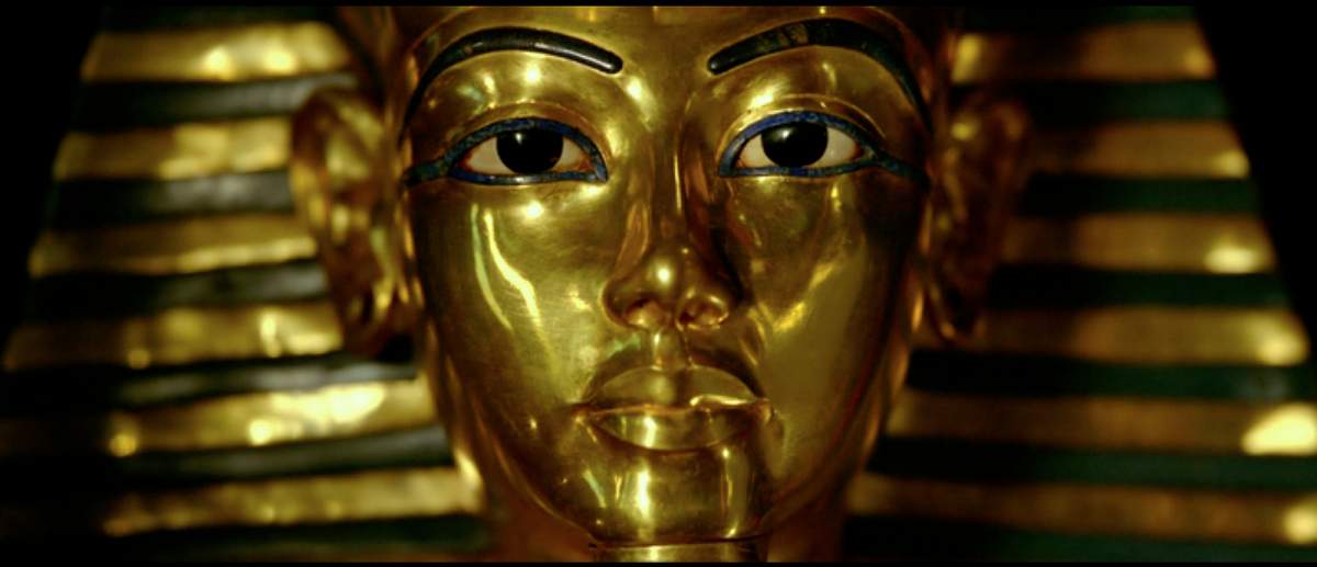 A close-up image of King Tutankhamen's burial mask. It is made out of gold. The only features on the face that are not gold are two thin black eyebrows and two large eyes with iris and pupils colored black.