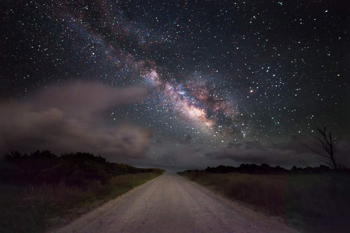 An image of a long dirt road. Above it is the night sky. On the left large white clouds are visible. On the right, the thousands of stars in the milky way.
