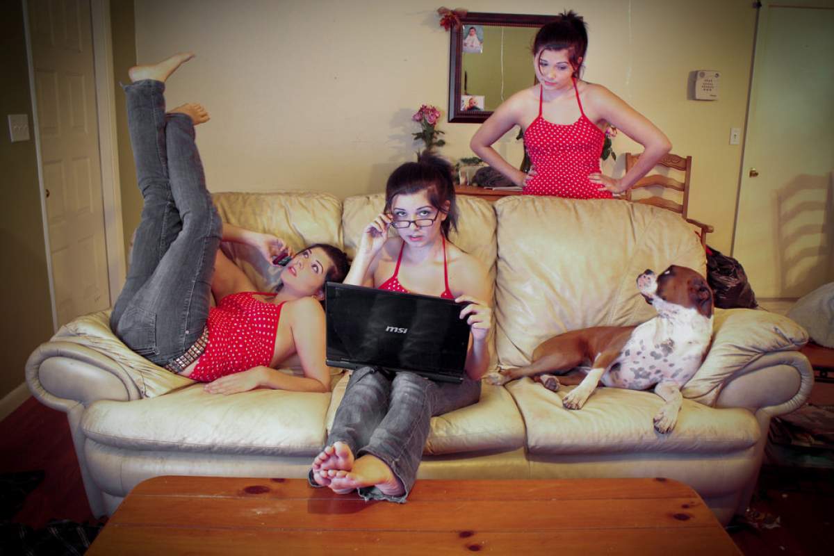A young woman sits on the couch with her legs in the air, next to a young woman who looks identical to her on a computer, and a brown and white dog sits next to that young woman. Behind the couch, another young woman, identical to the other two young women looks at the dog.