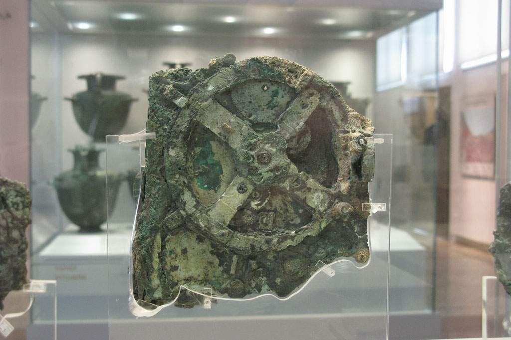 An image of the antikythera mechanism. It's enclosed in clear glass. The mechanism is green, light green, and dark colored stone.