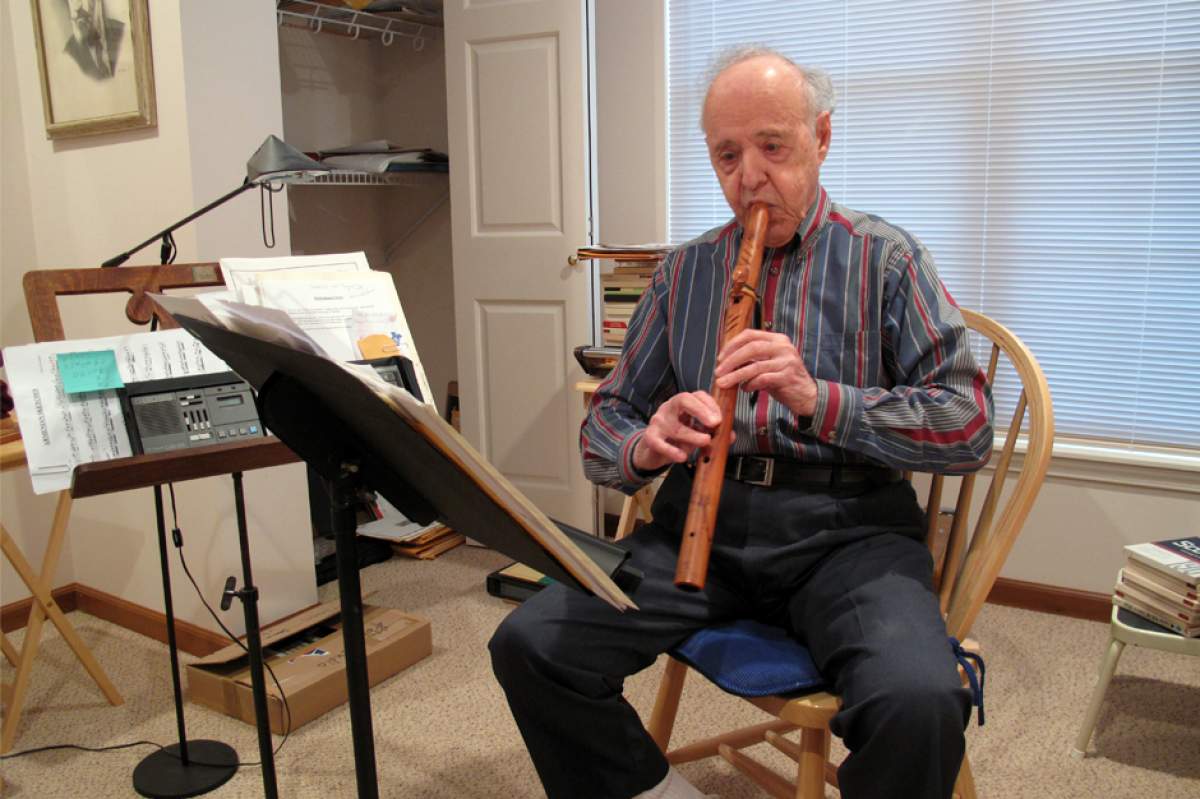 James Pellerite in his home practice studio, sitting in a chair, playing a wooden flute, looking at sheet music on a music stand