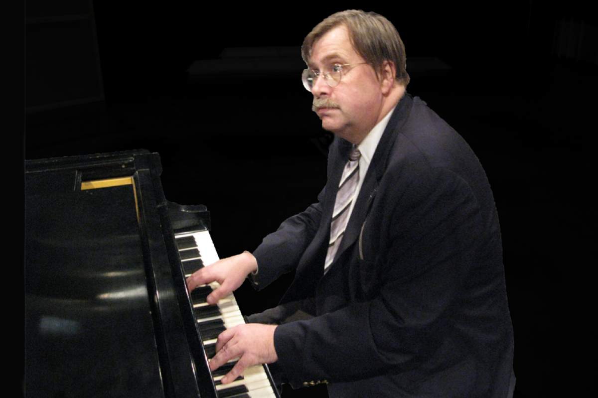 Philip Carli in a suit, sitting at a grand piano, looking up at the screen as he plays