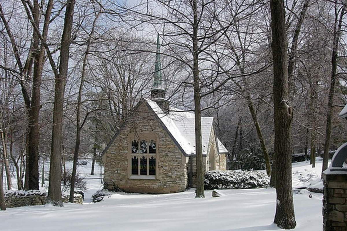 A small stone chapel in a wooded area is covered with a light snowfall.