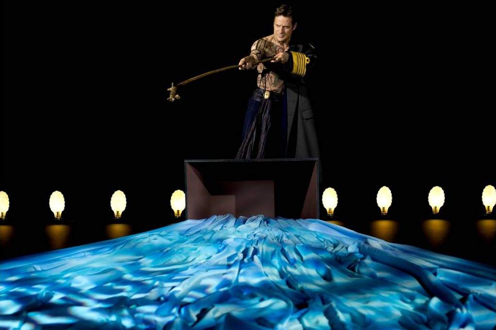 On stage, a tattooed man holds a wand over a shadowy pool of simulated water
