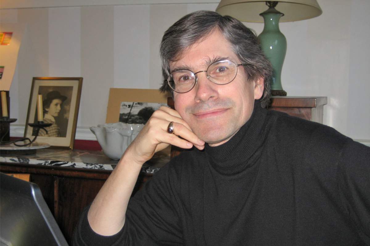 Ian Woollen sitting at a desk, in wireframe glasses and black turtleneck sweater, right hand on chin, smiling.