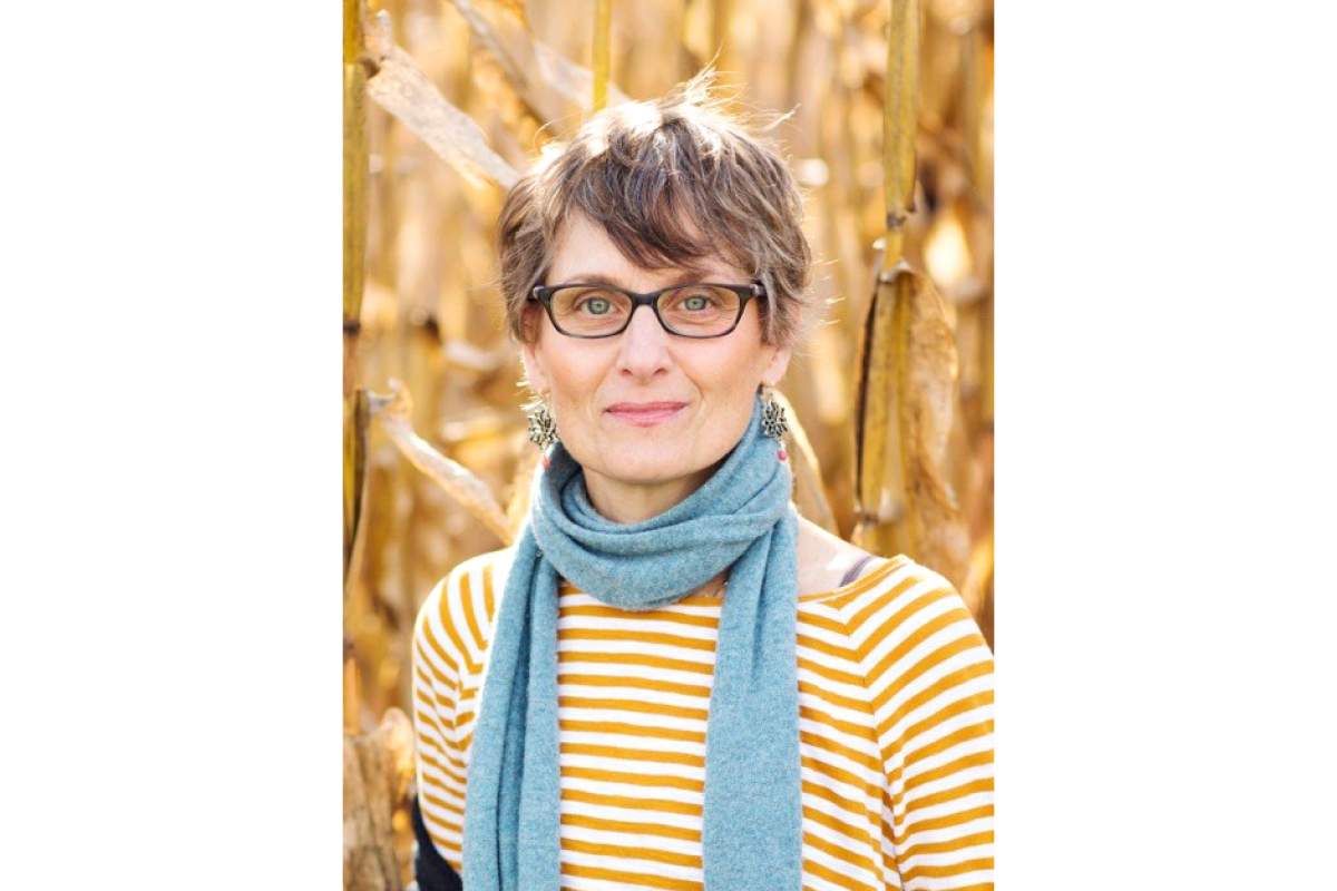 Liza Gennaro in cornfield, wearing horn-rimmed glasses, earrings, horizontal striped orange and white shirt, and blue scarf