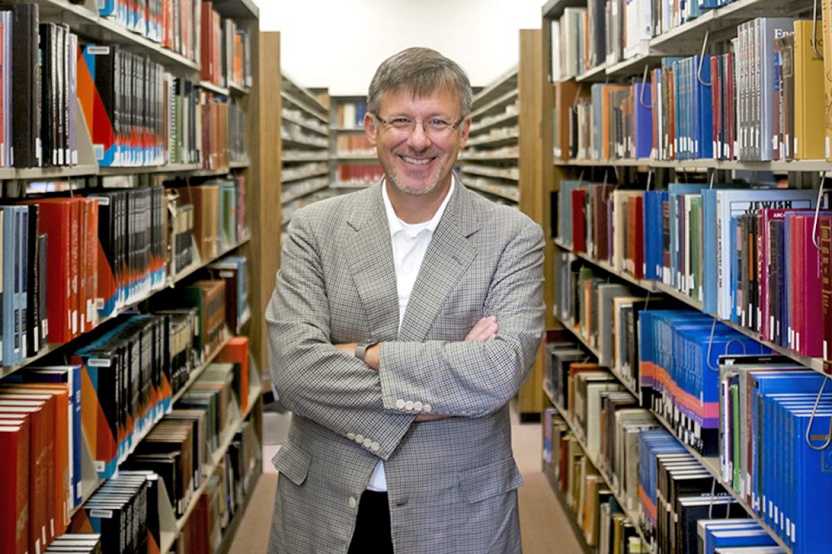 Michael Adams in glasses, black-and-white checked jacket over white dress shirt open at the collar, poses smiling and with arms crossed between stacks of books in the reference section of the Herman B Wells Library on Tuesday, July 26, 2016.