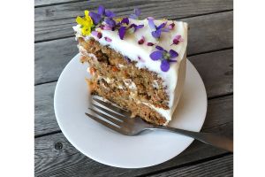 A slice of carrot cake, two layers with white icing and small purple and yellow flowers on top. the cake is standing up on a small white plate with a fork next to it. 