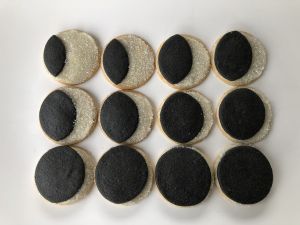 Twelve round, black and white cookies in a grid on a white plate. the decoration mimics the phases of a solar eclipse