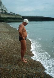 Doc, in his swim trunks, cap, and goggles, stands at water's edge on a cloudy day.