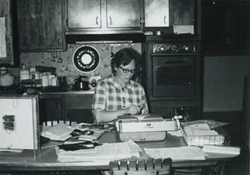 Marj works at a typewriter on the kitchen table, surrounded by notes and papers.