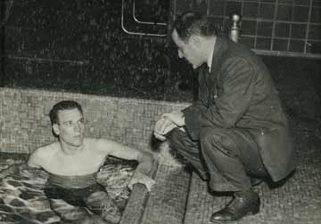 James, in the water at the edge of an indoor pool, talks to Coach Peppe, who kneels beside him.