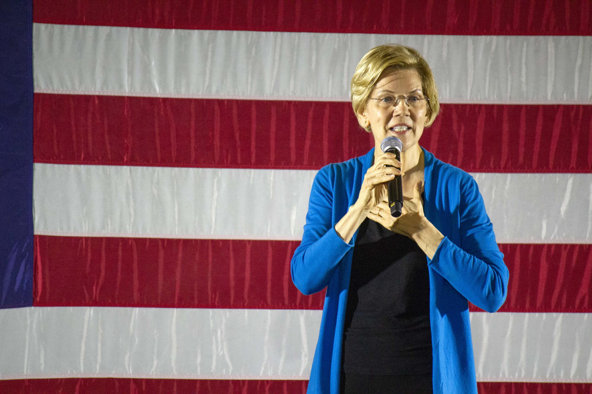 Elizabeth Warren standing in front of a large flag, mic in hand, hand on heart.