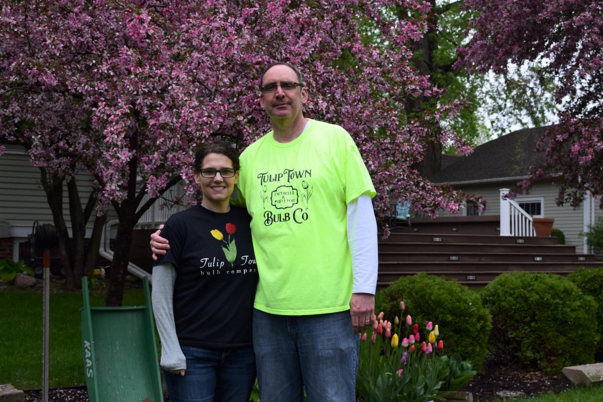 A tall man and a short woman stand in front of a tree in bloom with purple flowers. They both are wearing t-shirts that say "Tulip Town Bulb Co."