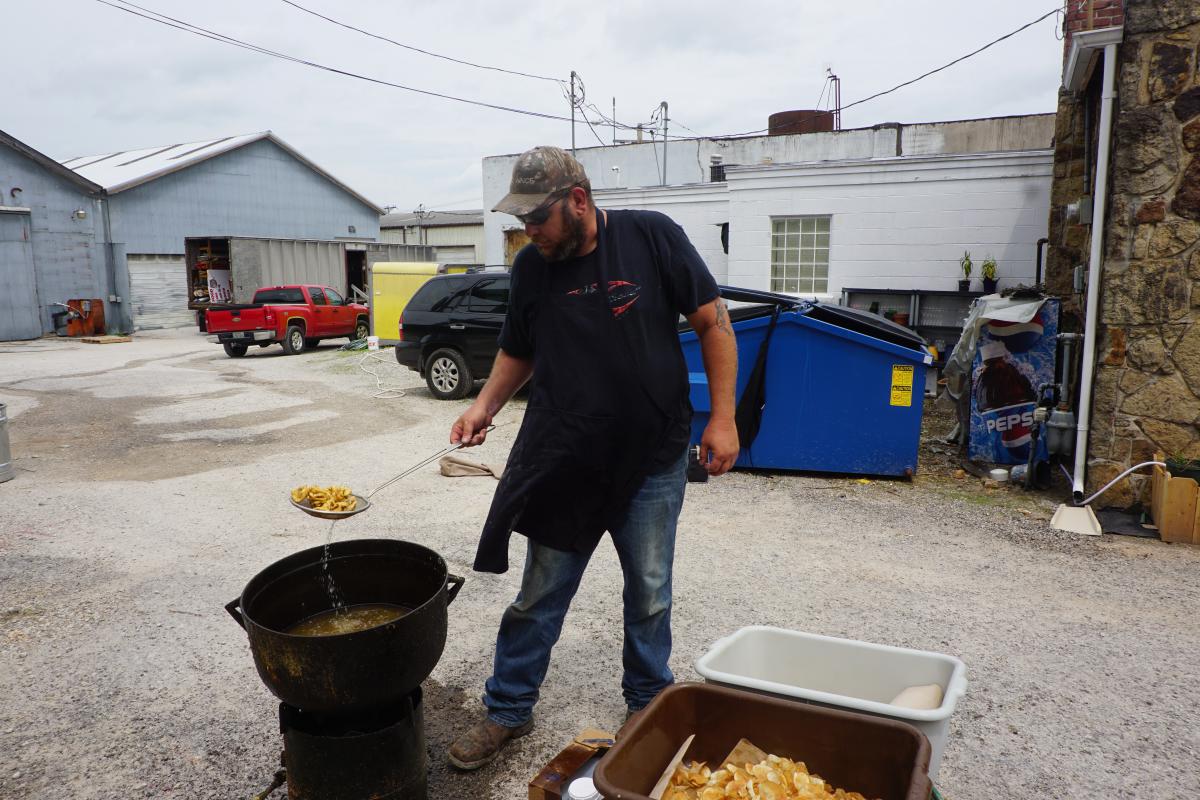 Bearded guy with hat, apron and sunglasses lifting fried fish from a cast iron kettle outdoors