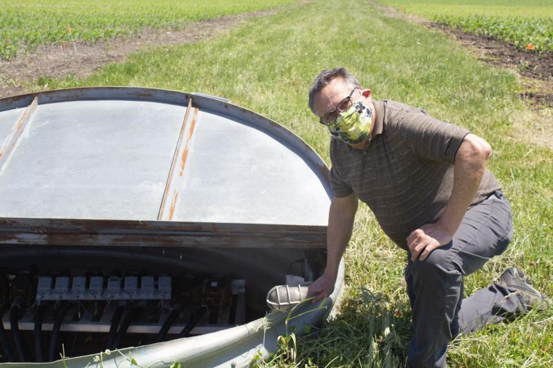 Tom Moorman, wearing a face mask, kneels beside a metal lined pit in a green field.