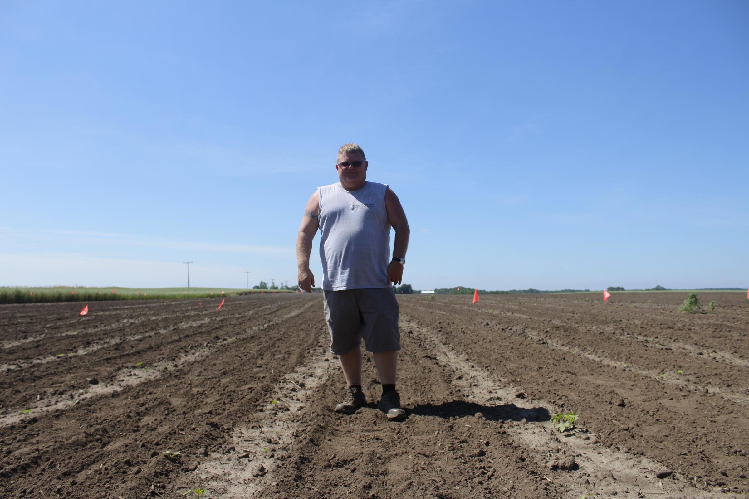 A big white guy in a sleeveless t-shirt standing in the middle of a plowed field with blue sky.