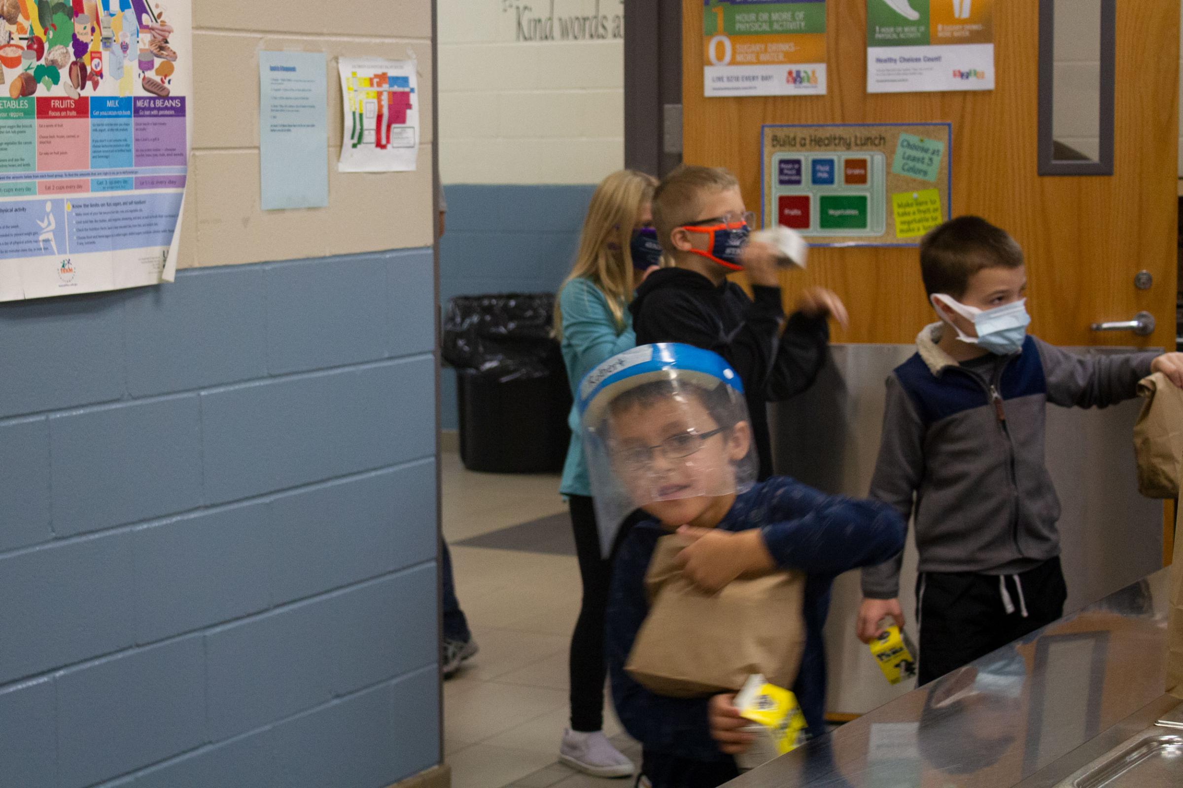 Kids in face masks and face shields move through a doorway with lunches and milk cartons in hand.