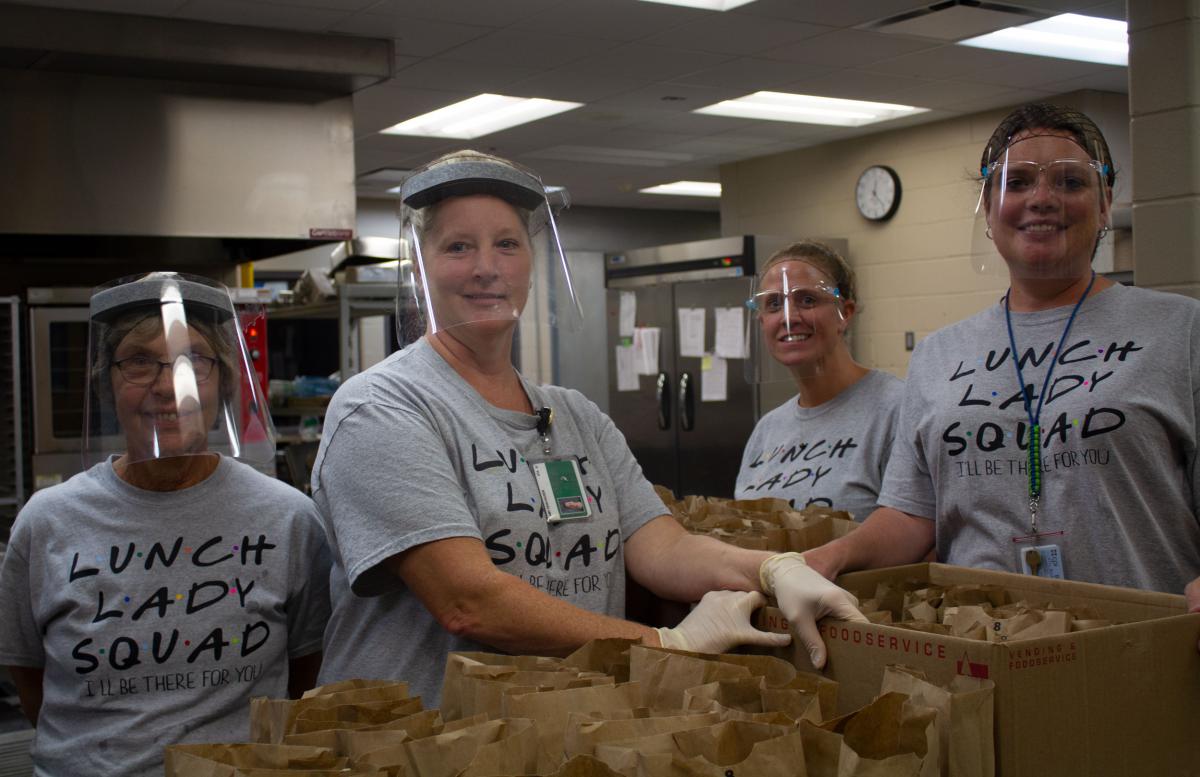 Four women in gray 'Lunch Lady Squad' t-shirts and clear face shields stand with filled brown paper bags in front of them in an institutional kitchen.