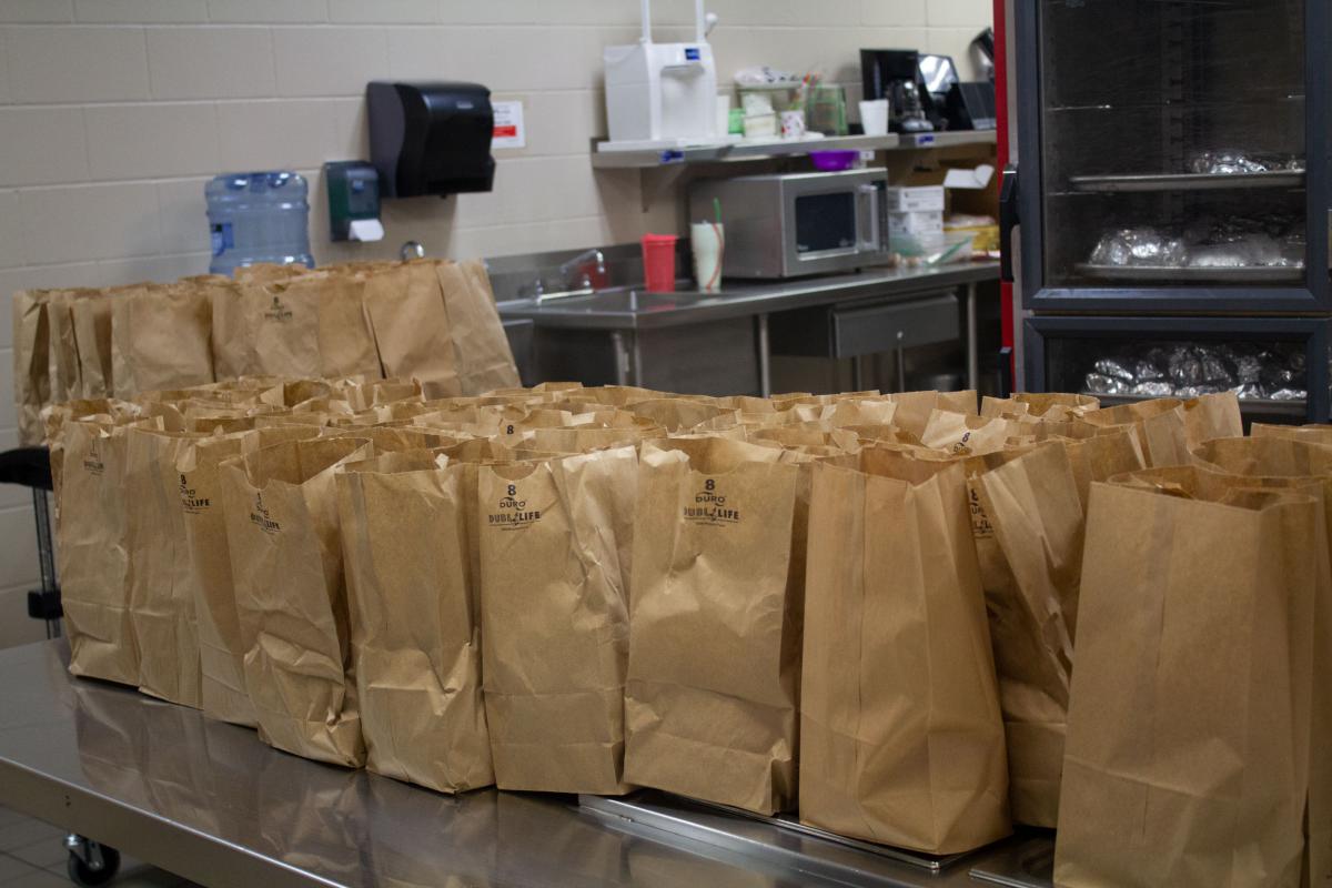 Rows of filled brown paper bags line a stainless steel table in a commercial kitchen.