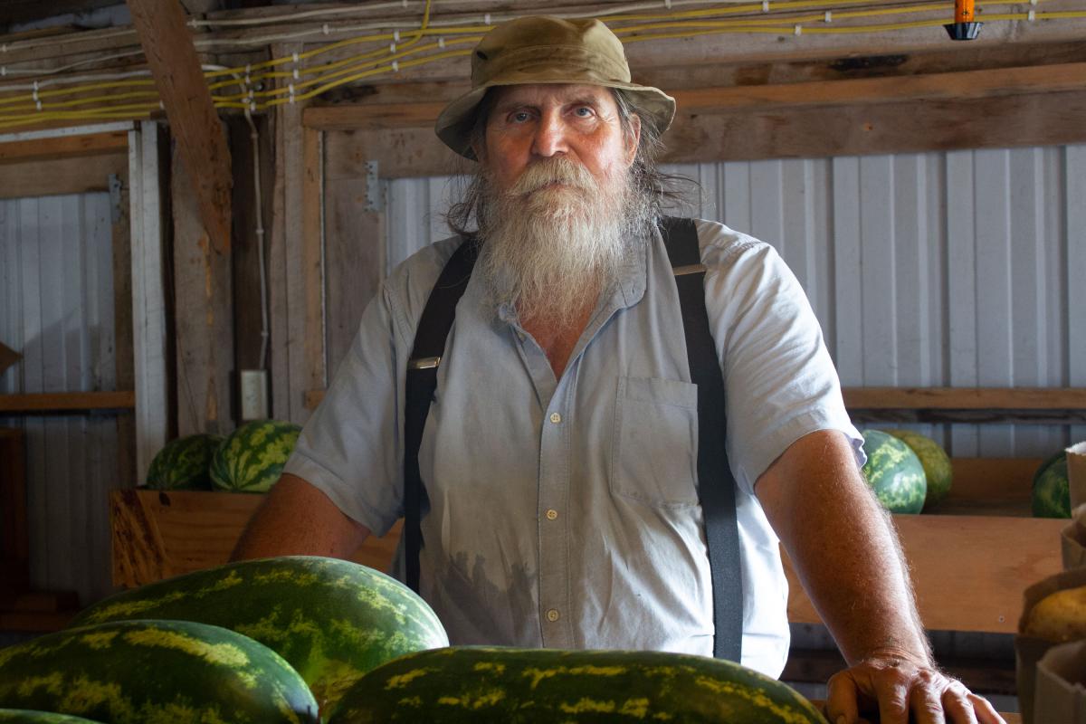 Charles Downs with hat and suspenders stands with some watermelons inside a barn..