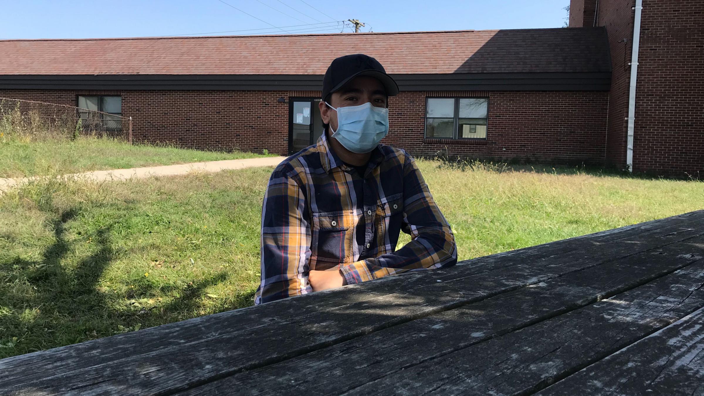 Samuel Gomez sitting at a wooden table in the shade with a brick building behind him. 