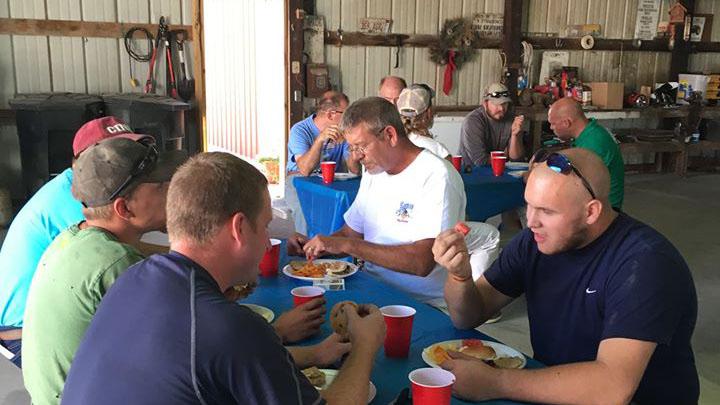 Men (mostly caucasian) in t-shirts sitting around tables eating inside a metal sided barn.