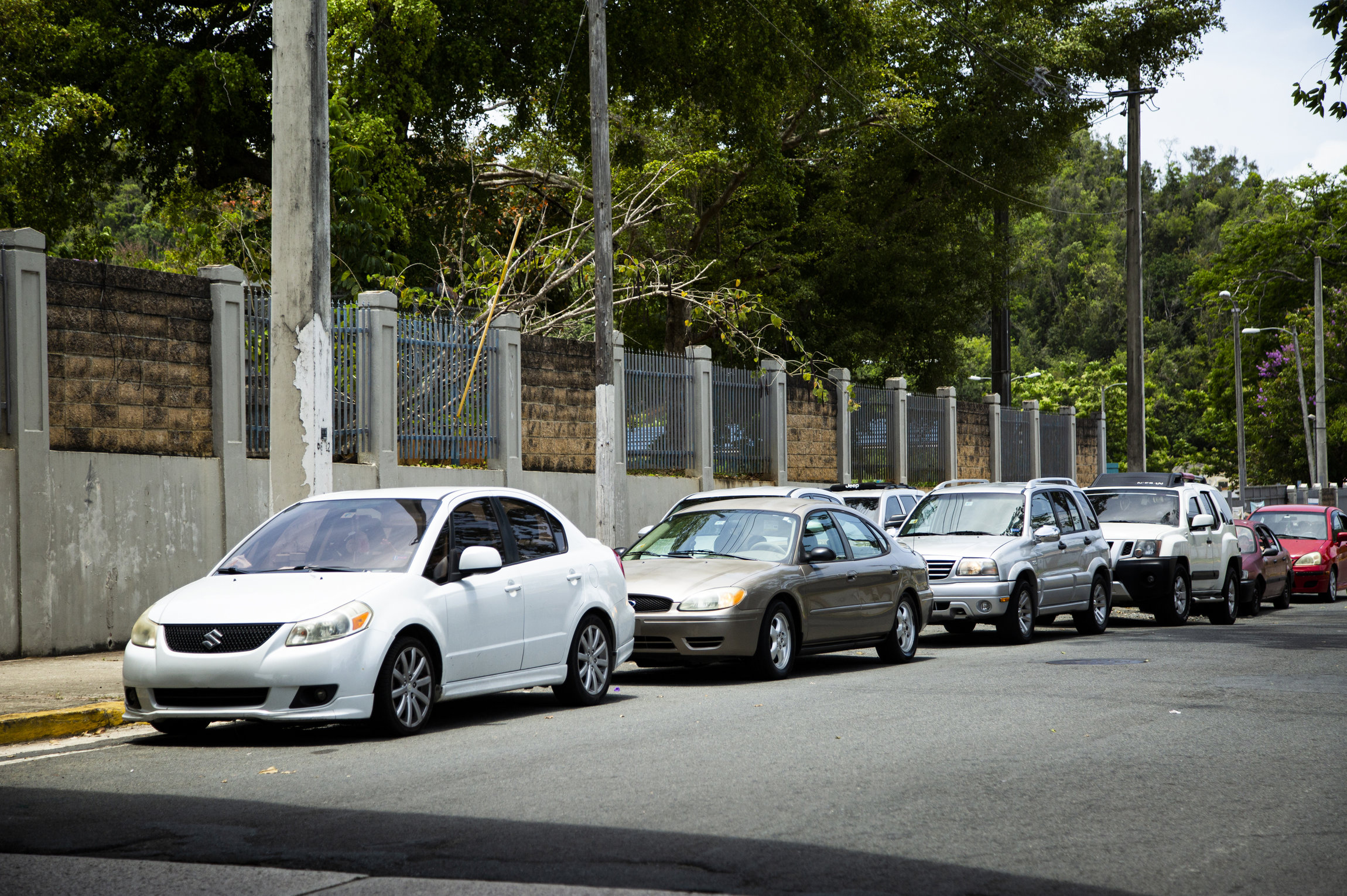 A line of cars, bumper to bumber, on a road next to a wall. Behind the wall is lush vegetation.