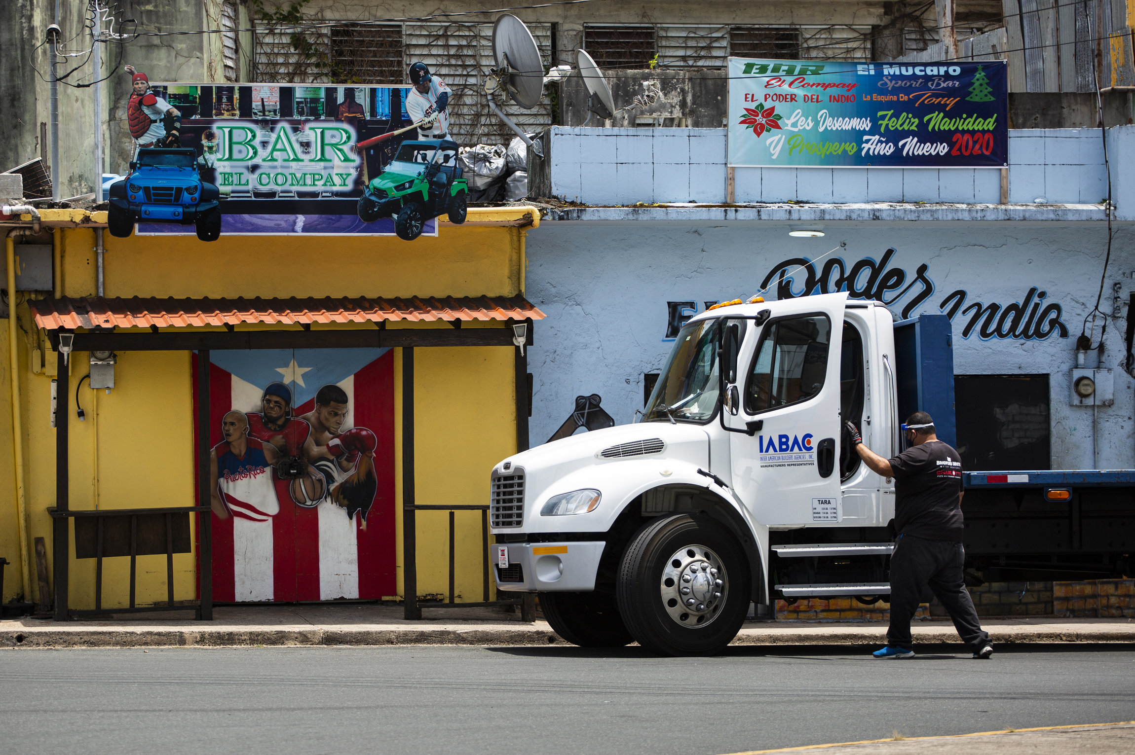 A man with a face shield opens the door to a white truck cab in front of a bar with images of sports figures and trucks. 