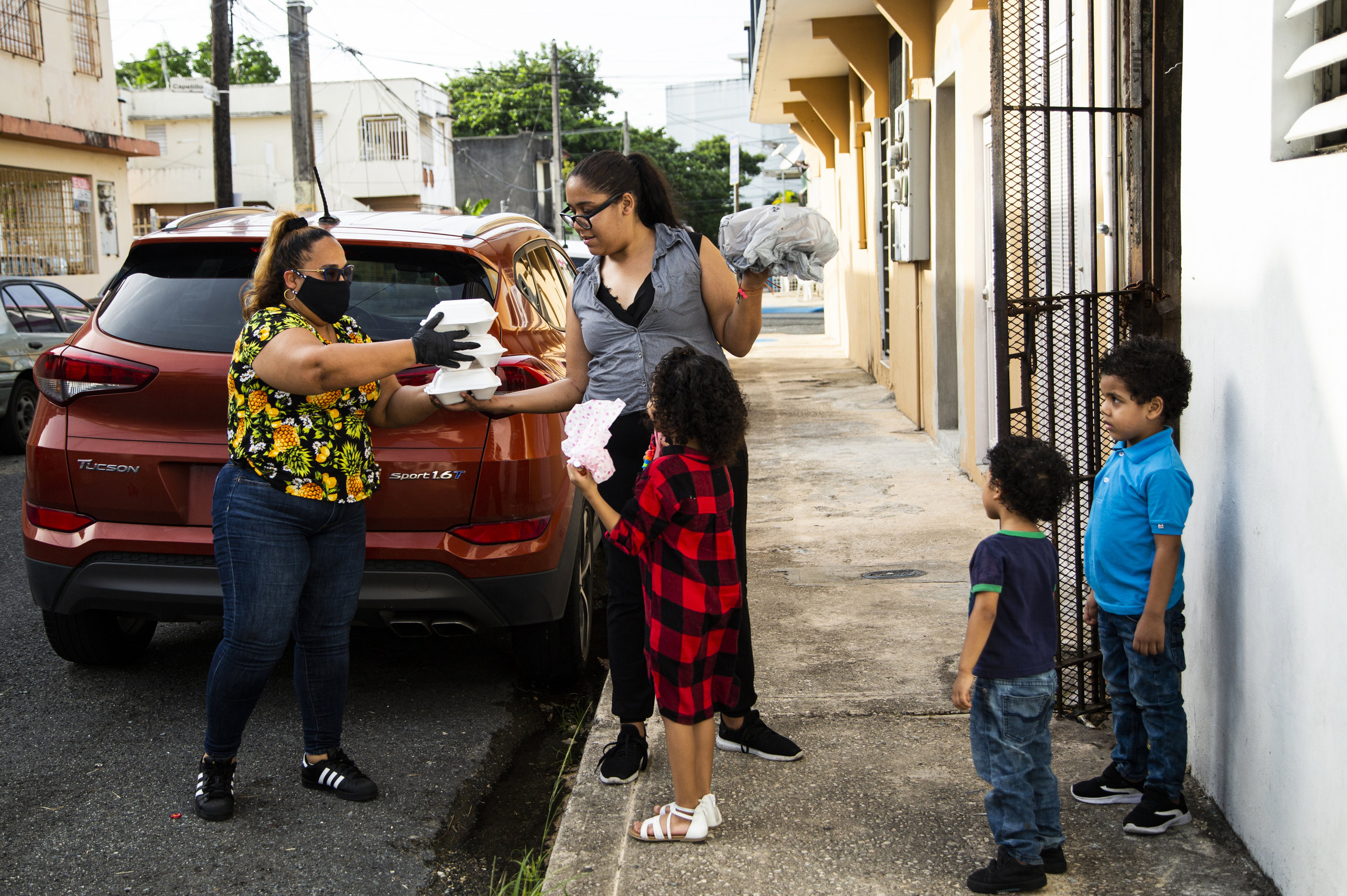 Christel Galindez Garcia, in a mask and gloves, passes a stack of white clamshell food containers to Elia Gonzalez, who stands next to her three young children, next to a parked car and near a doorway.