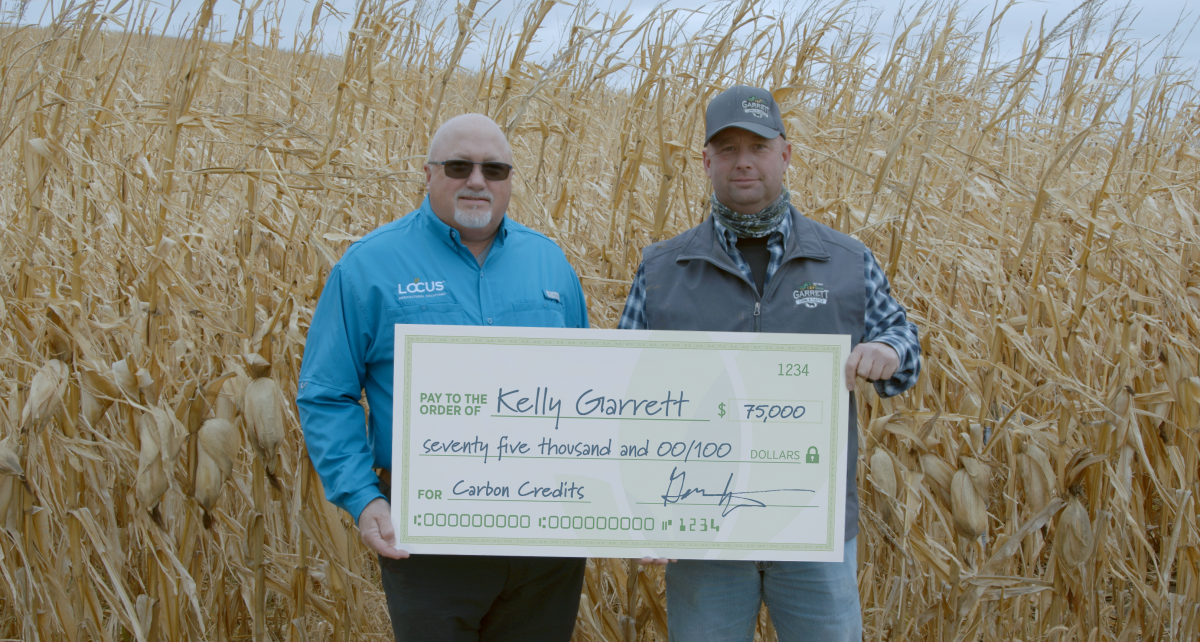 Kelly Garrett and Shane Head standing in front of a field of dry corn holding a giant check for $75,000 with "cabon credits" in the memo line
