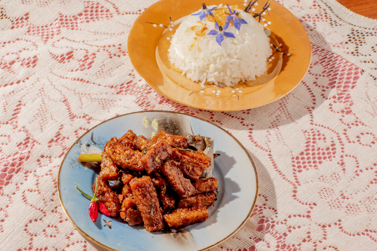 A plate with a rounded serving of rice decorated with flowers and another plate with strips of tempeh in a red sauce all arranged on a lace background