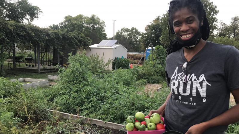 Ja Nelle Pleasure, smiling, with a face mask at her chin, stands in a garden near tomato plants holding a bowl of green and red tomatoes.