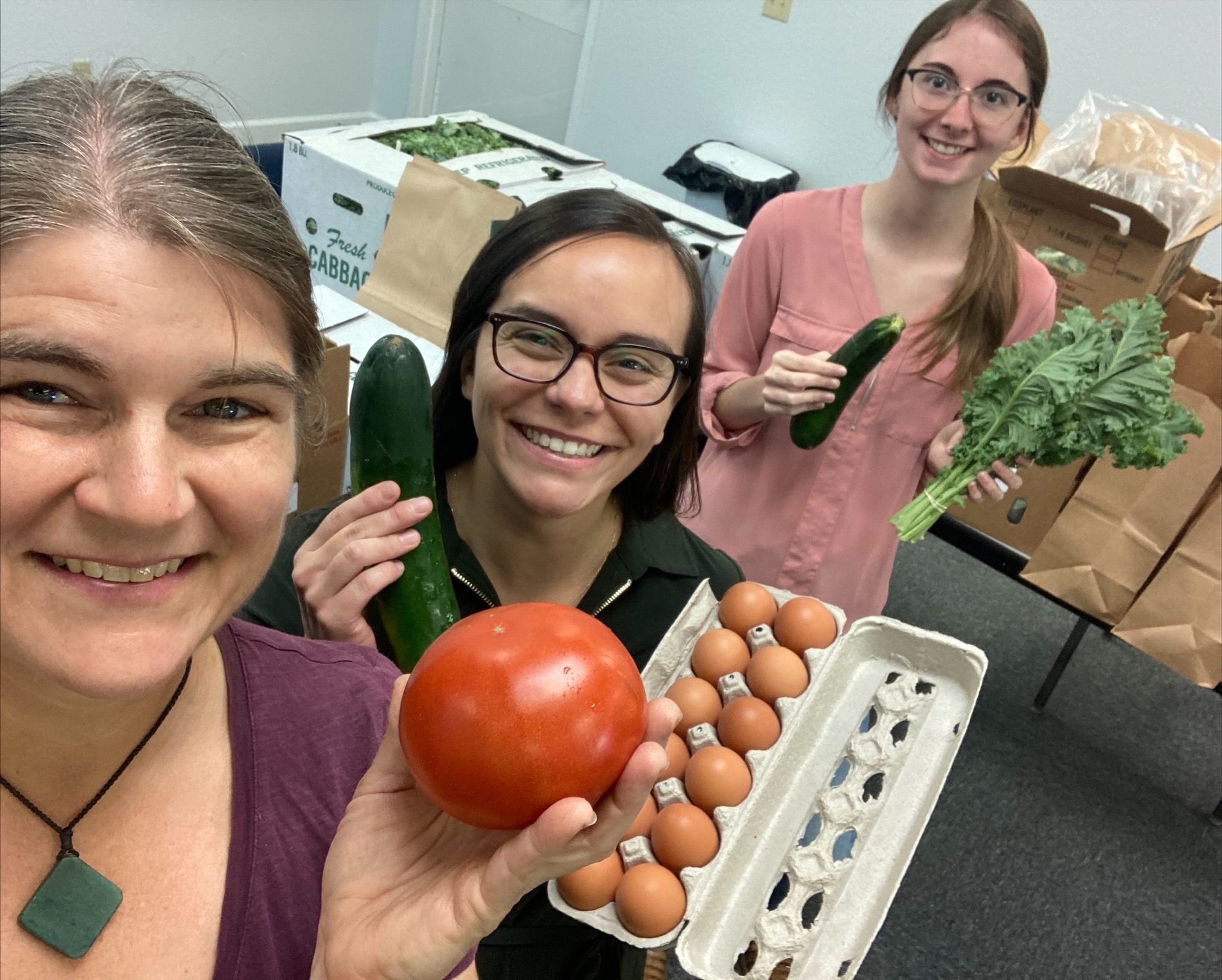 three women smiling at camera holding tomato, carton of brown eggs and a bunch of greens