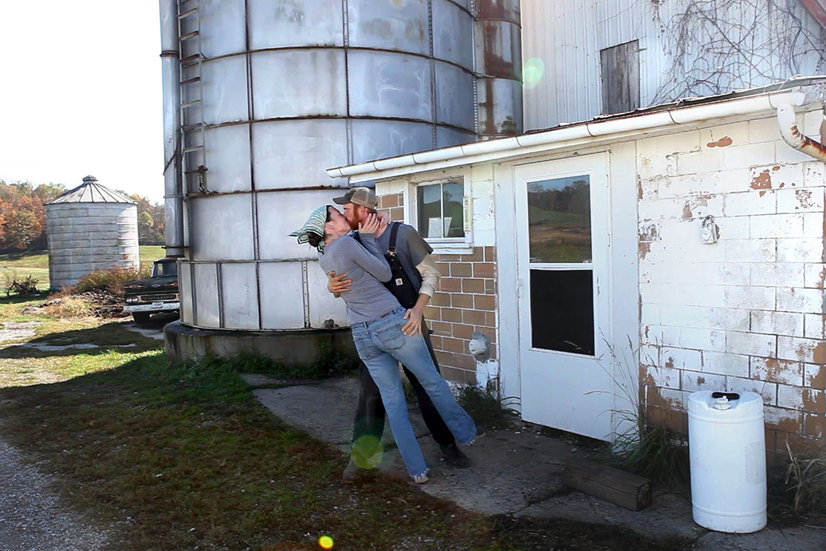 A man in overalls dipping and kissing a woman in jeans, in front of an old farm building with silos and rural landscape in the background.