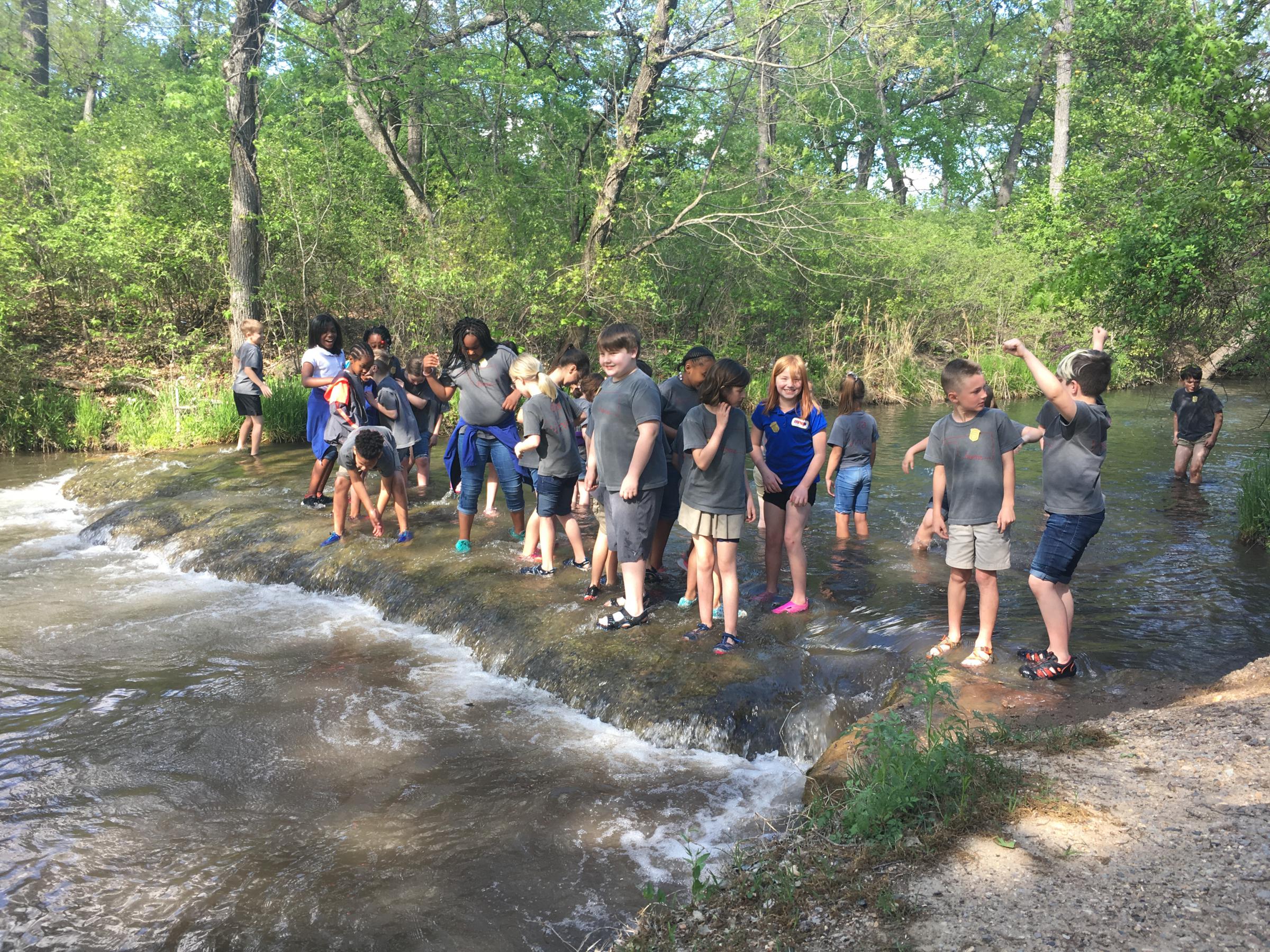 A large group of school-aged kids in gray t-shirts and shortsstanding in a wide shallow creek, smiling and laughing.