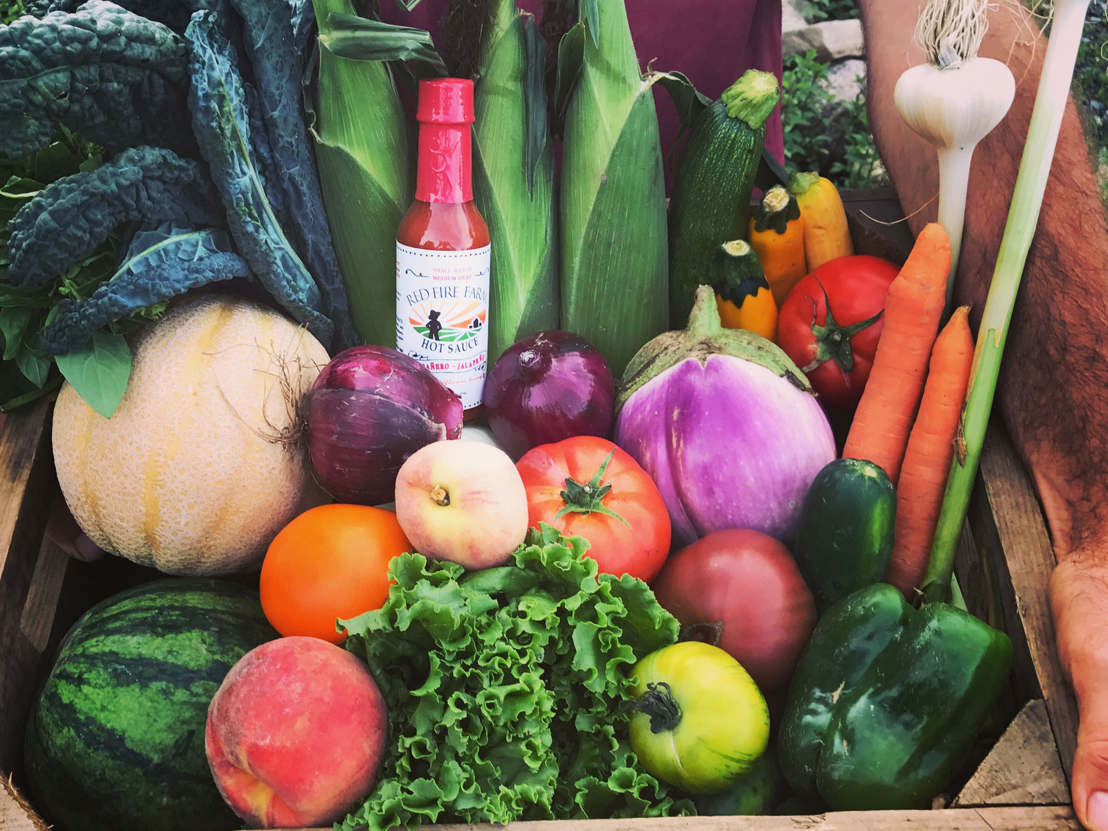 A wooden crate filled with colorful farm fresh produce and a bottle of hotsauce.