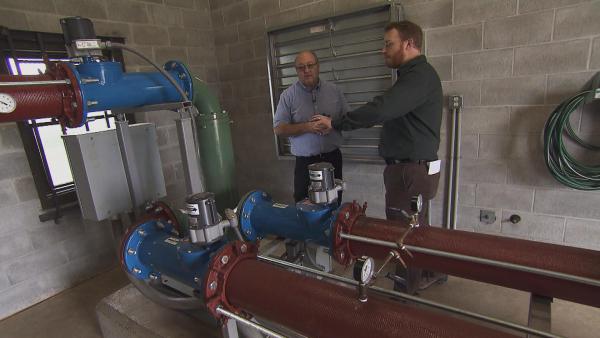 Marty Stange talks with another man next to large pipes, guages and valves in a cinderblock-walled room