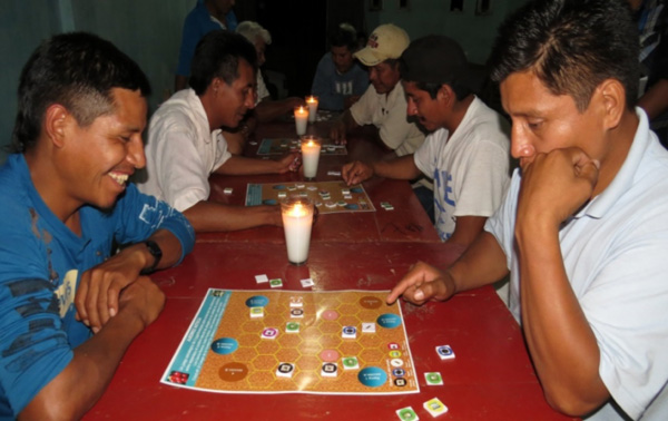 Two men sitting across from each other with a board game between them, and a candle with others in the background