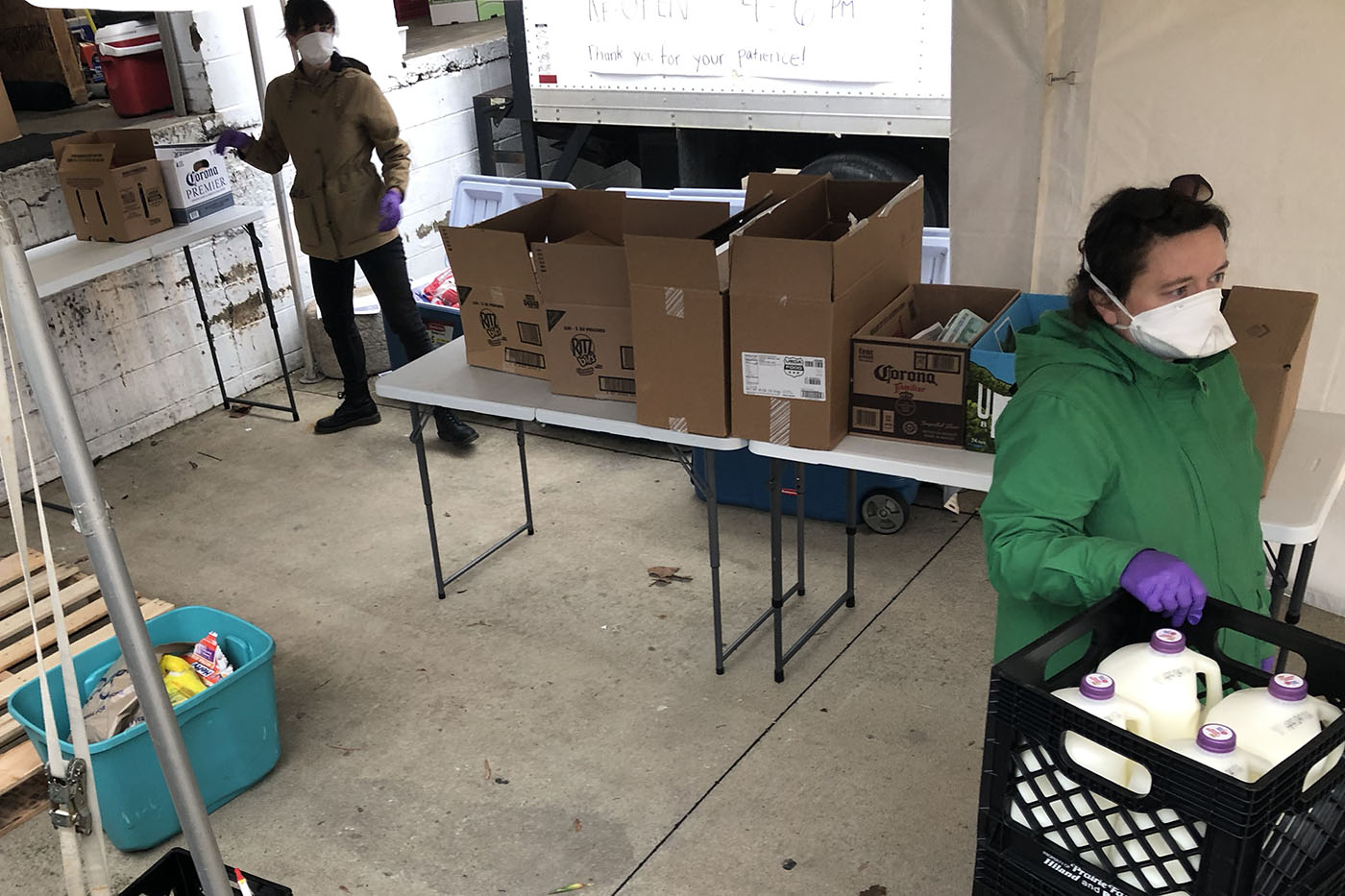 Two women in masks and gloves, outside in a truck bay under a white party tent with a table full of food boxes and a stack of milk crates with milk. 