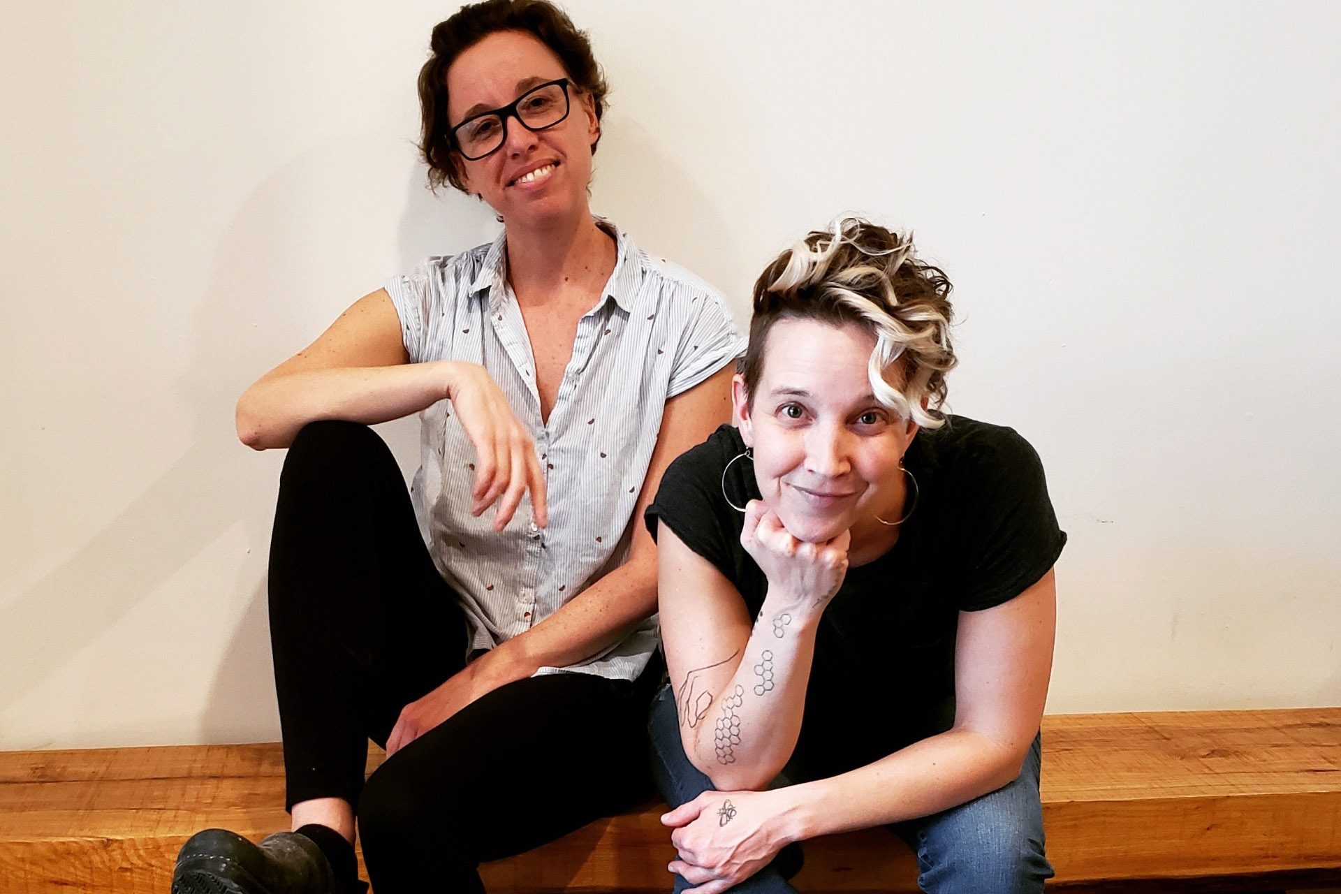 Kassie Jensen and Amanda Armstron, sitting on the wooden bench in their bakery. Kassie leaning against white wall, Amanda leaning forward with chin resting on hand.