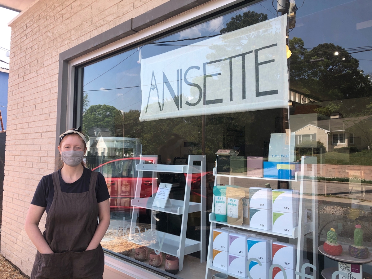 Nicole Evans Groth in apron with face covering,  standing in front of a plate glass window with display shelves and and Anisette sign visible