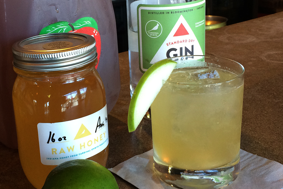 Light green cocktail with a thin apple slice on rim, with spirit bottles in the background.