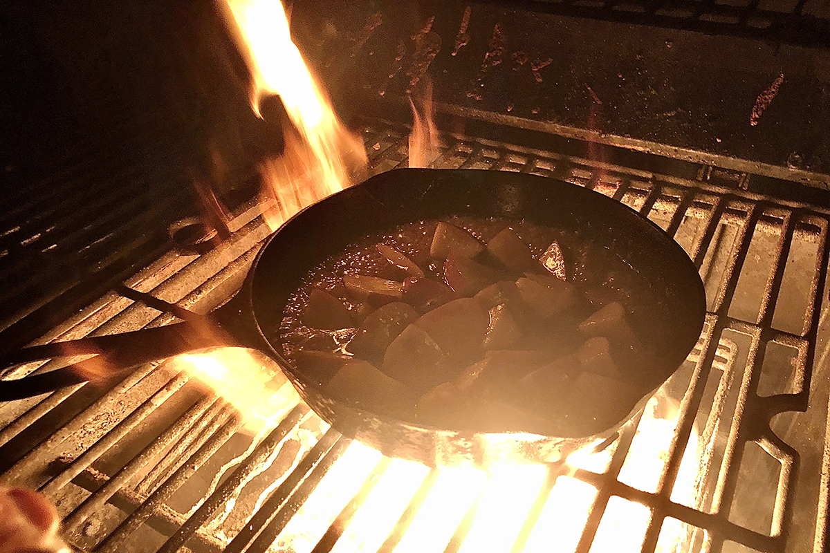 A cast iron skillet with apple slices, bubbling on a grill with fire beneath it, in the dark with natural light 