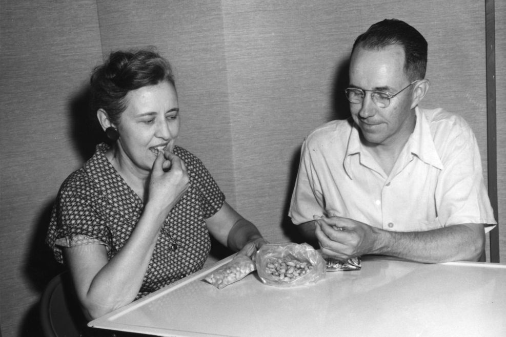 Black and white photo of man and woman tating peanuts from a bag, at a table.