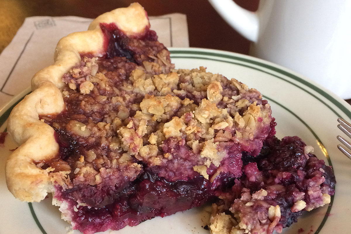 Slice of pie with purple berries and a crumble topping on a small plate with coffee cup in the background.