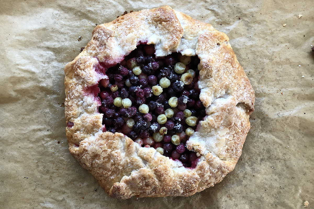 Rustic baked tart , in a rough, cirular shape, with purple and green berries in the middle, on brown parchment paper.