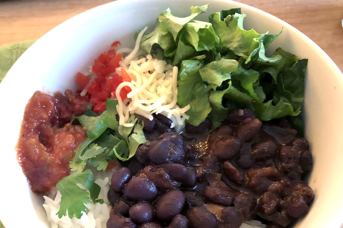 A white bowl with cooked black beans topped with cheese, lettuce, salsa, cilantro.