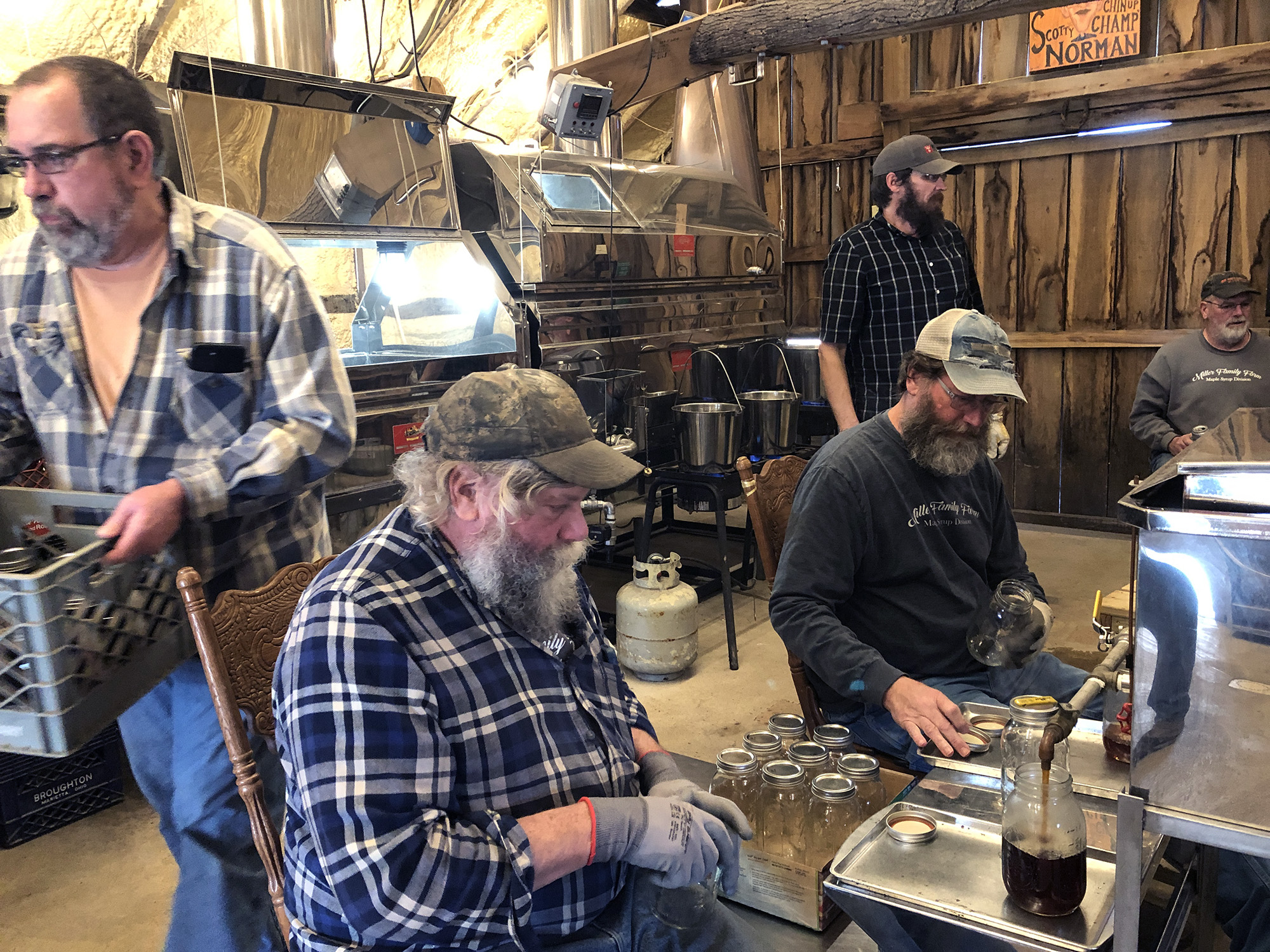 several white men with beards in a barn-type room with equipment, and a station where brown liquid is going into jars