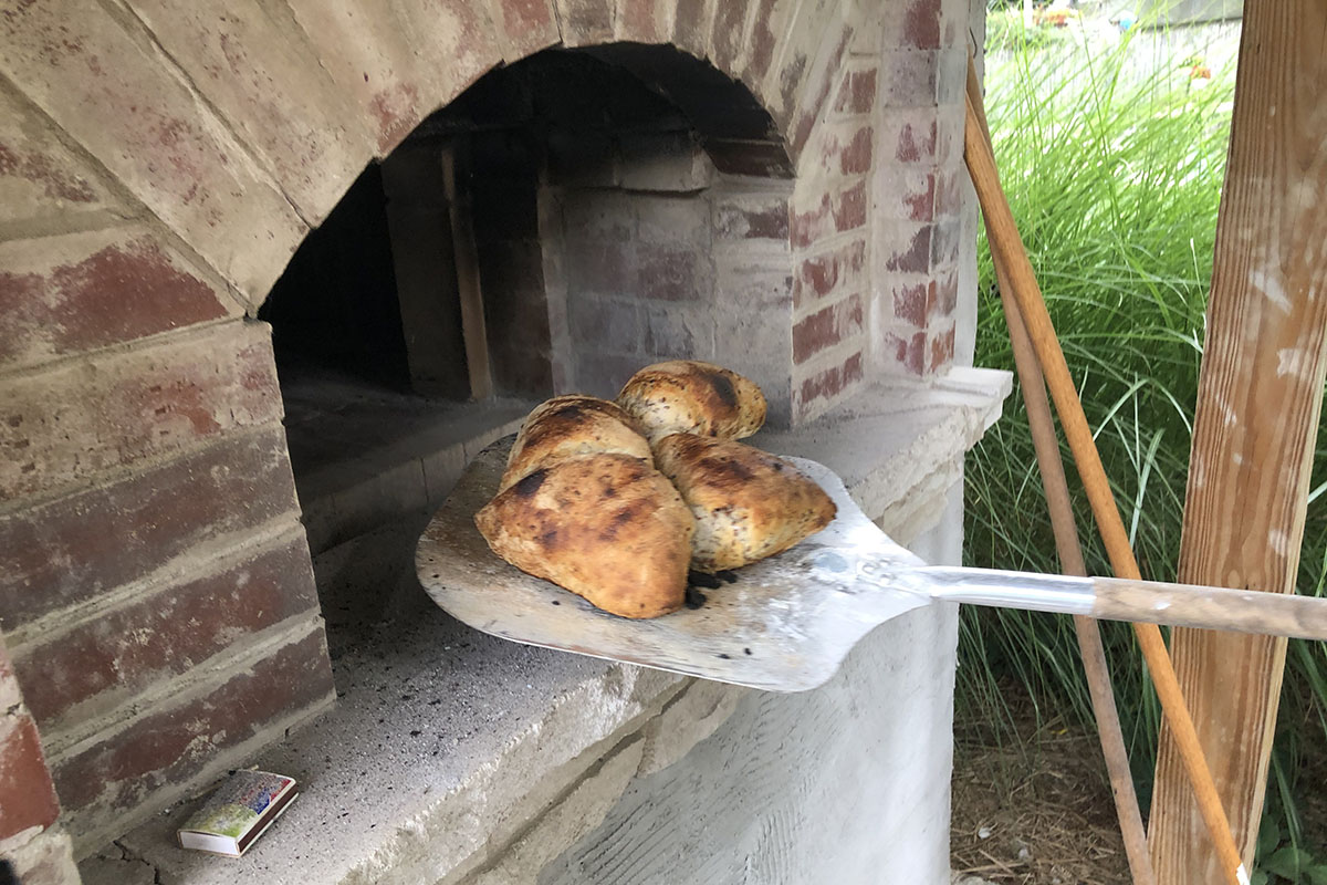 Four uniquely shaped loaves of bread on a metal pizza peel, at the entrance of a brick oven. 